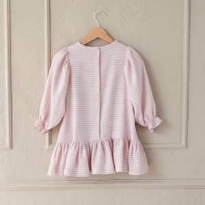 May include: A light pink and white gingham dress with long sleeves and a ruffled hem, hanging on a wooden hanger. The dress has a round neckline and button closure at the back. The sleeves have a slight puff at the shoulder and are gathered at the cuffs.