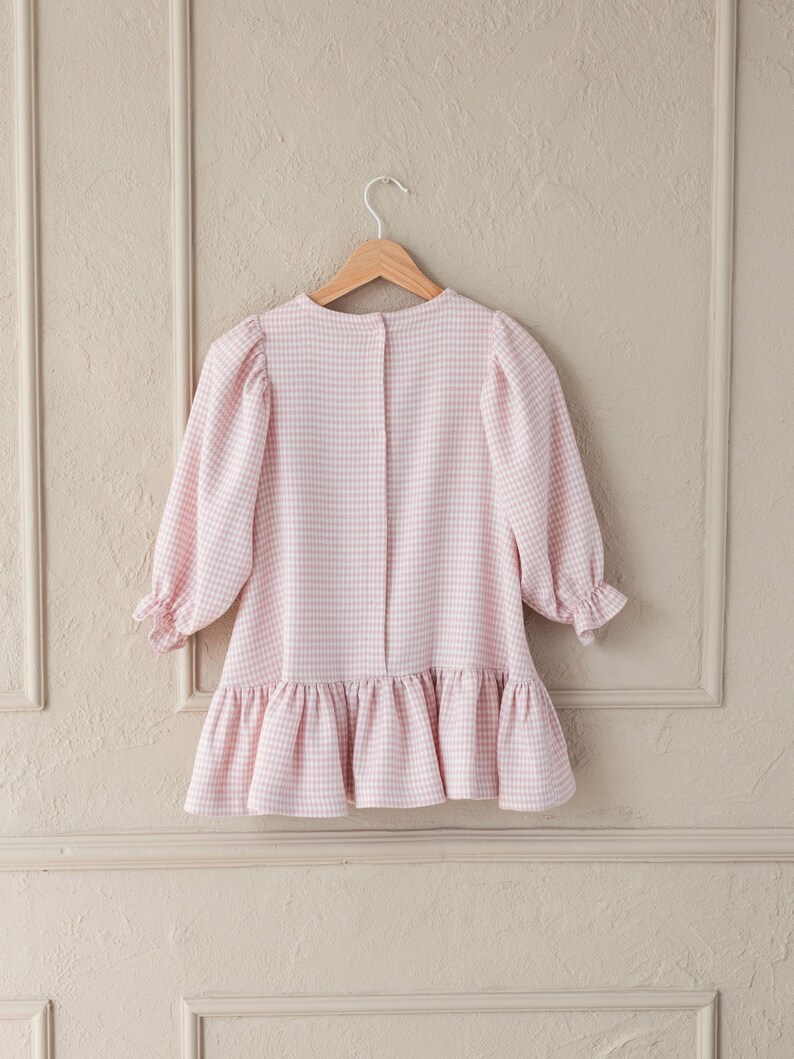 May include: A light pink and white gingham dress with long sleeves and a ruffled hem, hanging on a wooden hanger. The dress has a round neckline and button closure at the back. The sleeves have a slight puff at the shoulder and are gathered at the cuffs.