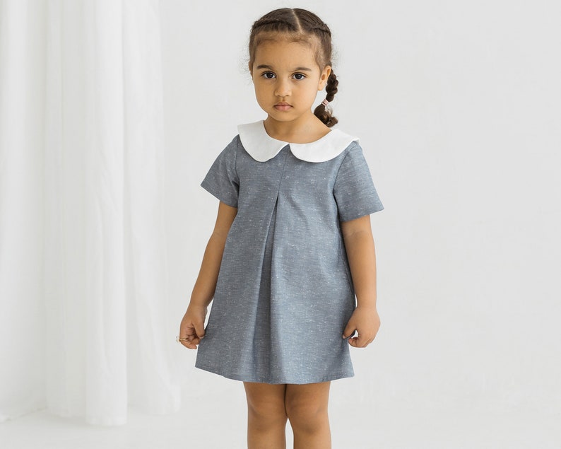 May include: A young girl wearing a gray dress with a white Peter Pan collar. The dress has a pleated skirt and short sleeves.