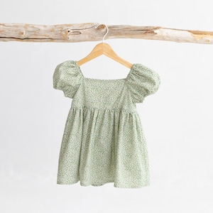 Puede incluir: Un vestido de estampado floral verde y blanco con mangas abullonadas y escote cuadrado. El vestido est&aacute; colgado en una percha de madera.