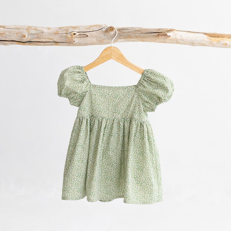Puede incluir: Un vestido de estampado floral verde y blanco con mangas abullonadas y escote cuadrado. El vestido est&aacute; colgado en una percha de madera.