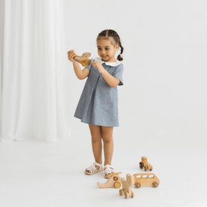May include: A young girl in a light blue dress with a white collar plays with wooden toys. The toys include a small airplane, a bus, and an elephant.