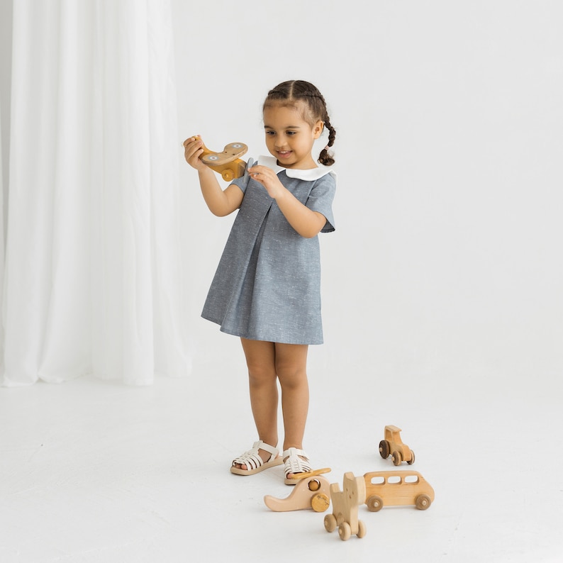 May include: A young girl in a light blue dress with a white collar plays with wooden toys. The toys include a small airplane, a bus, and an elephant.