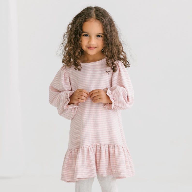 May include: A light pink and white gingham dress with long, puffy sleeves and a ruffled hem. The dress is worn by a young child with curly brown hair, and is paired with white leggings. The dress has a round neckline.