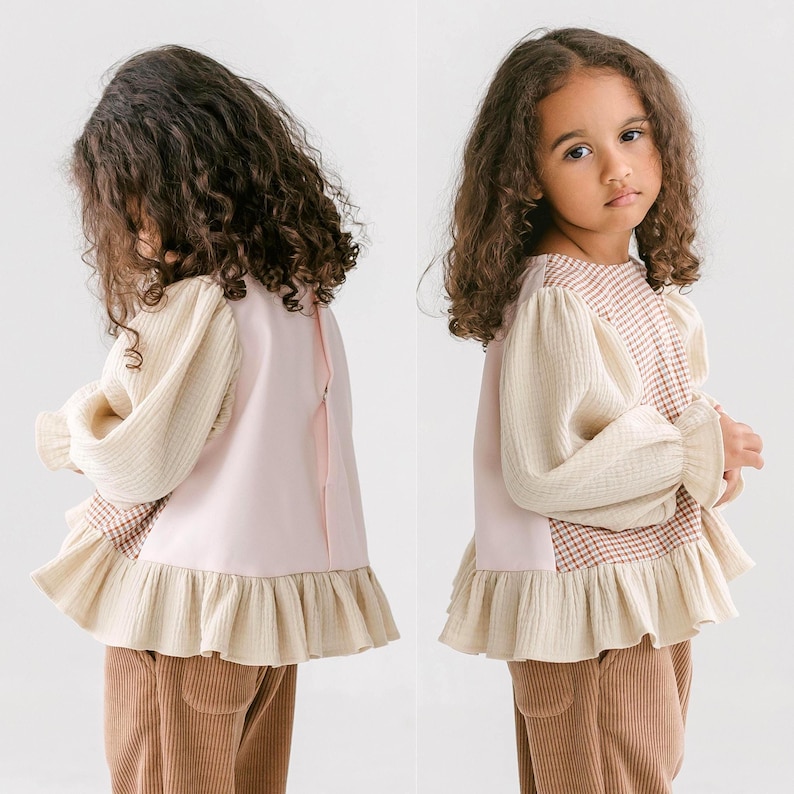 May include: A light pink and cream-colored top with a ruffled hem and sleeves. The top features a plaid pattern on the upper chest and cream-colored sleeves. The model is wearing brown corduroy pants.