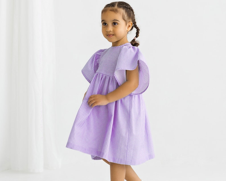 May include: A young girl wearing a purple and white gingham dress with puffed sleeves. The dress has a gathered skirt and a round neckline.