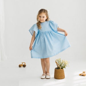 May include: A young girl wearing a light blue gingham dress with puffed sleeves. She is standing in a white room with a wooden toy car and a wicker basket with flowers.