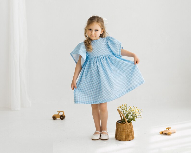 May include: A young girl wearing a light blue gingham dress with puffed sleeves. She is standing in a white room with a wooden toy car and a wicker basket with flowers.