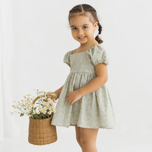 Puede incluir: Una ni&ntilde;a peque&ntilde;a con un vestido floral verde y blanco con mangas abullonadas. Est&aacute; sosteniendo una cesta de mimbre llena de flores blancas.