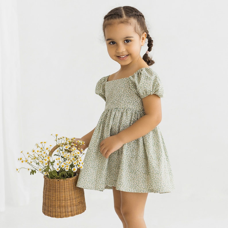 Puede incluir: Una ni&ntilde;a peque&ntilde;a con un vestido floral verde y blanco con mangas abullonadas. Est&aacute; sosteniendo una cesta de mimbre llena de flores blancas.