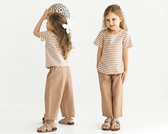 May include: Two young girls wearing brown and white striped shirts and brown linen pants. One girl is holding a leopard print ball and the other is standing with her hands in her pockets.