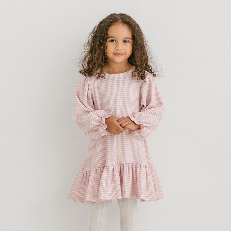May include: A light pink gingham dress with long, puffy sleeves and a ruffled hem. The dress is worn by a young person with curly brown hair. The dress is a classic style, suitable for casual or semi-formal occasions.