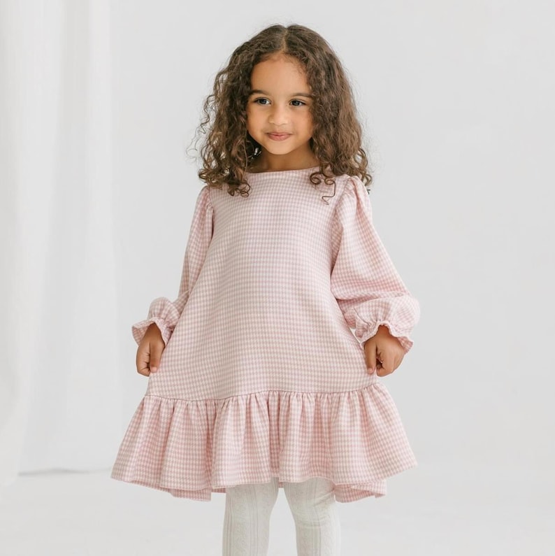 May include: A light pink and white gingham dress with long sleeves and a ruffled hem. The dress has a round neckline and is worn with white leggings. The sleeves have ruffled cuffs. The dress is worn by a young child.