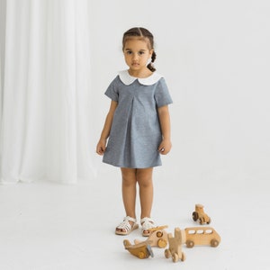 May include: A young girl wearing a light blue dress with a white collar stands in front of a white background. She is playing with wooden toy cars and airplanes.