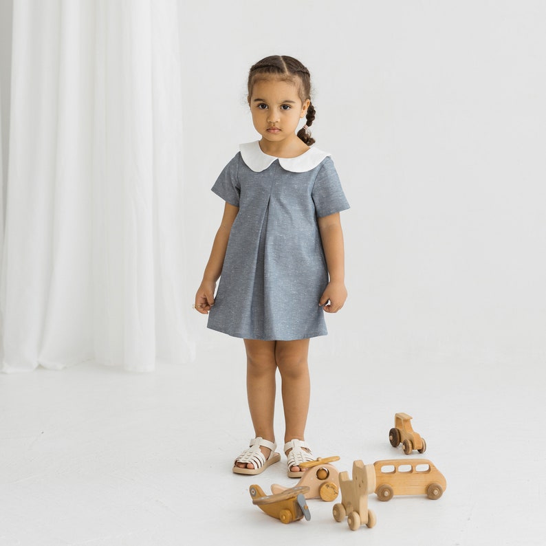 May include: A young girl wearing a light blue dress with a white collar stands in front of a white background. She is playing with wooden toy cars and airplanes.