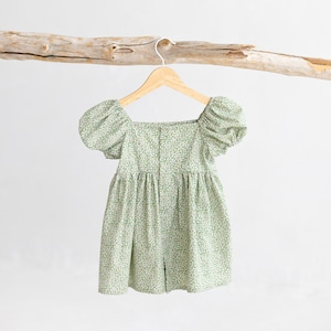 Puede incluir: Un vestido de estampado floral verde y blanco con mangas abullonadas y escote cuadrado. El vestido est&aacute; colgado en una percha de madera.