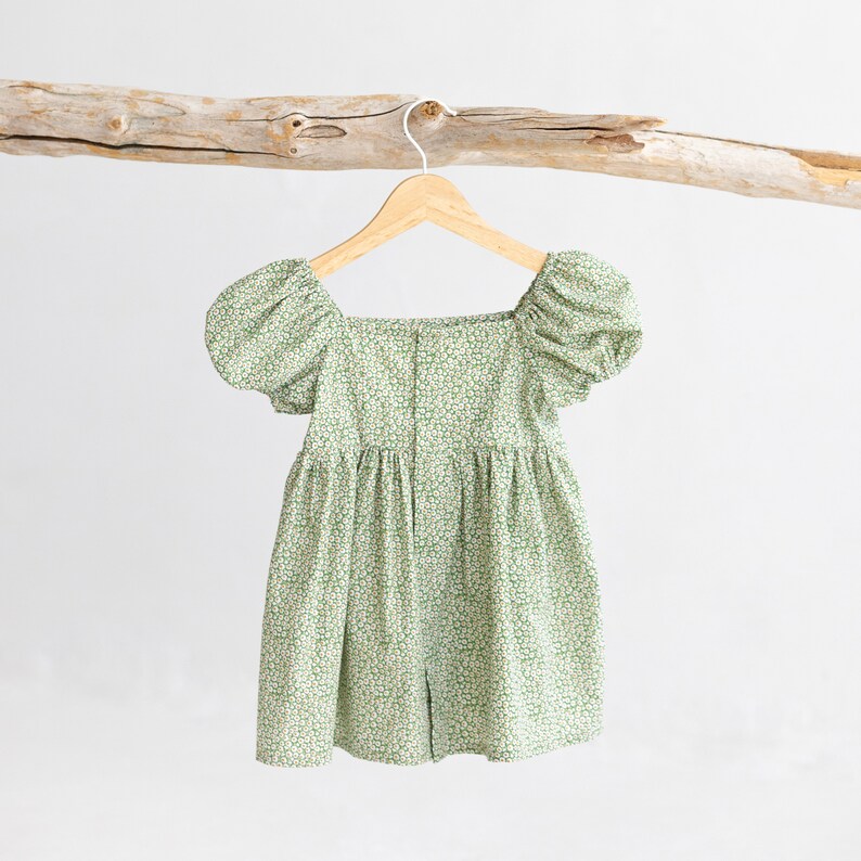 Puede incluir: Un vestido de estampado floral verde y blanco con mangas abullonadas y escote cuadrado. El vestido est&aacute; colgado en una percha de madera.