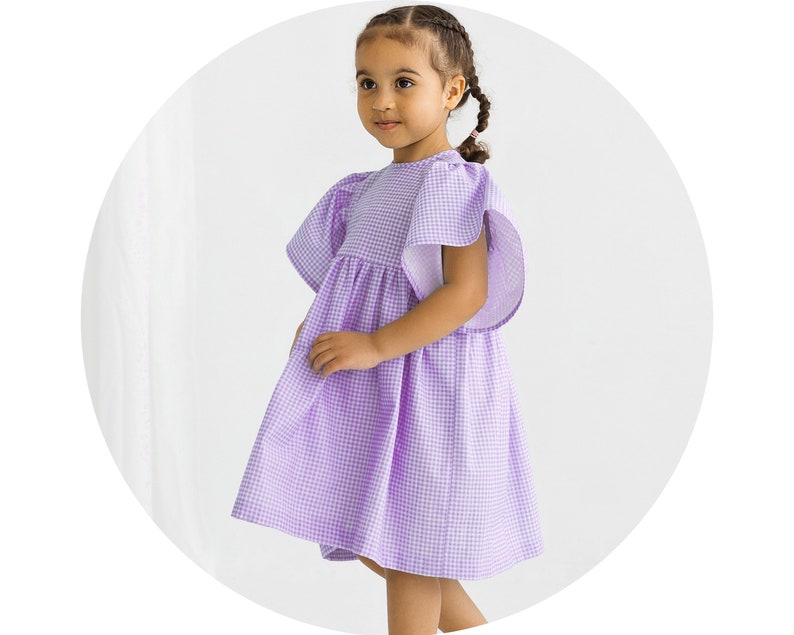 May include: A young girl wearing a purple and white gingham dress with short puffed sleeves. The dress has a gathered skirt and a round neckline.