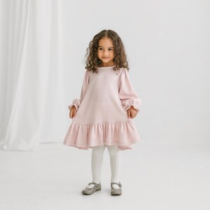 May include: A little girl wearing a pink and white checkered dress with long sleeves and a ruffled hem. She is also wearing white tights and gray Mary Jane shoes. The dress has a round neckline and a loose fit.
