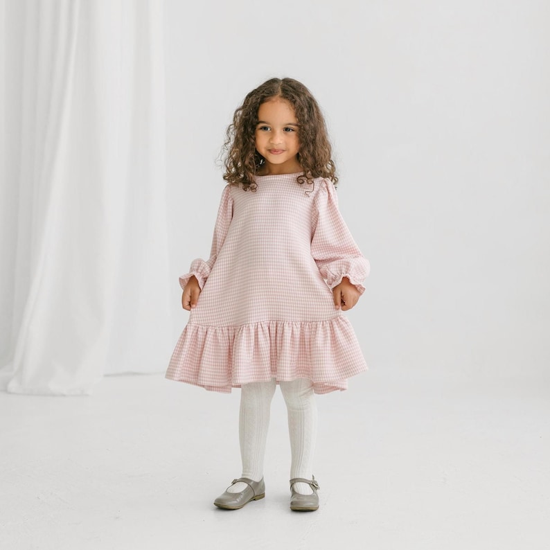 May include: A little girl wearing a pink and white checkered dress with long sleeves and a ruffled hem. She is also wearing white tights and gray Mary Jane shoes. The dress has a round neckline and a loose fit.
