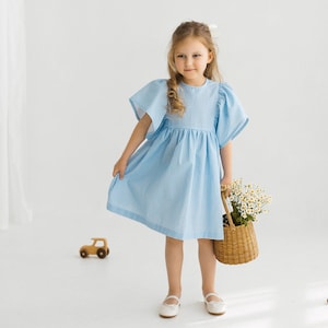 May include: A young girl in a light blue gingham dress holds a wicker basket filled with white flowers. She is standing on a white background.