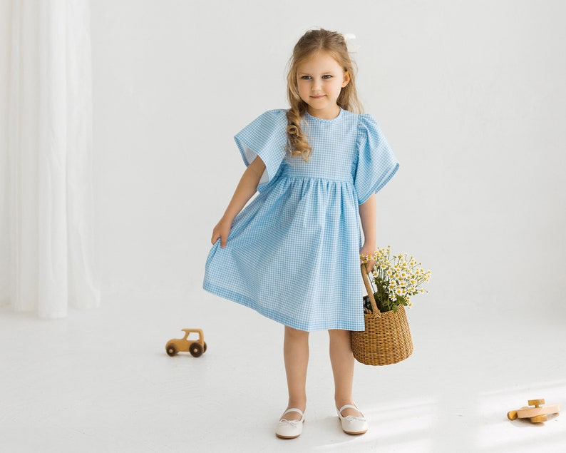 May include: A young girl in a light blue gingham dress holds a wicker basket filled with white flowers. She is standing on a white background.