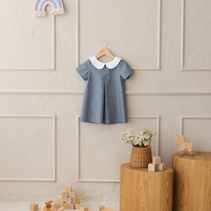 May include: A blue and white collared dress hangs on a wooden hanger against a light gray wall. The dress has a white collar and short sleeves. There are wooden blocks and toy vehicles on the floor in front of a wooden table with a wicker basket of flowers.