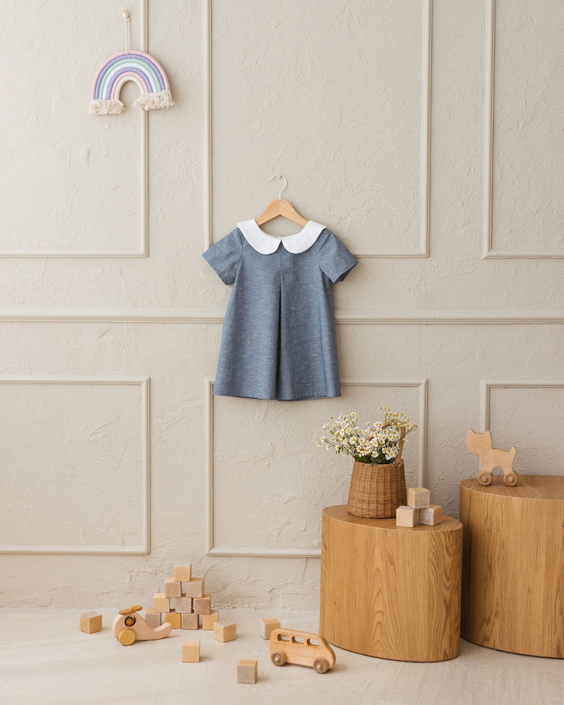 May include: A blue and white collared dress hangs on a wooden hanger against a light gray wall. The dress has a white collar and short sleeves. There are wooden blocks and toy vehicles on the floor in front of a wooden table with a wicker basket of flowers.