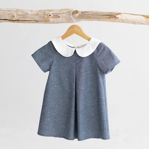 May include: A blue linen dress with a white peter pan collar and short sleeves. The dress is hanging on a wooden hanger.
