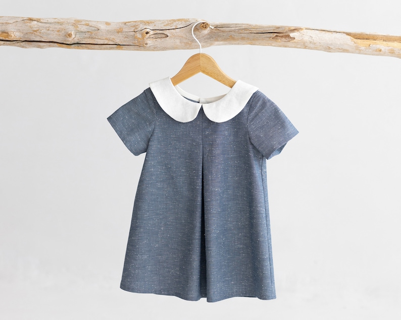 May include: A blue linen dress with a white peter pan collar and short sleeves. The dress is hanging on a wooden hanger.
