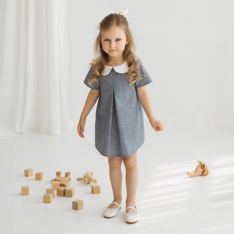 May include: A young girl wearing a gray dress with a white collar and white shoes. She is standing on a white floor with wooden blocks and a wooden toy car.