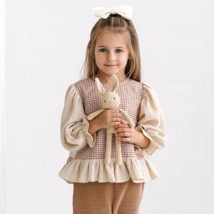 Peut inclure: Une jeune fille porte un haut à carreaux beige et rouge avec de longues manches à volants et un pantalon en velours côtelé marron. Elle tient un jouet lapin crocheté beige avec de longues oreilles. Un nœud blanc est dans ses cheveux.