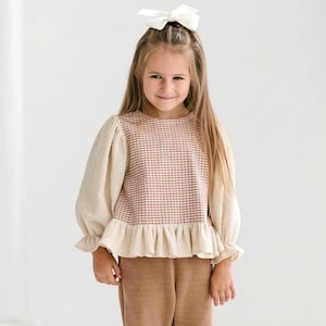 May include: A young girl wearing a beige and brown plaid top with long, puffy sleeves and a ruffled hem. She is also wearing brown corduroy pants and a white bow in her hair. The outfit is a mix of textures and patterns.