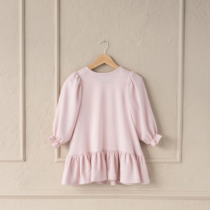 May include: A light pink and white gingham dress with long sleeves and a ruffled hem. The dress is hanging on a wooden hanger against a neutral background. The dress has a round neckline and puffed sleeves.