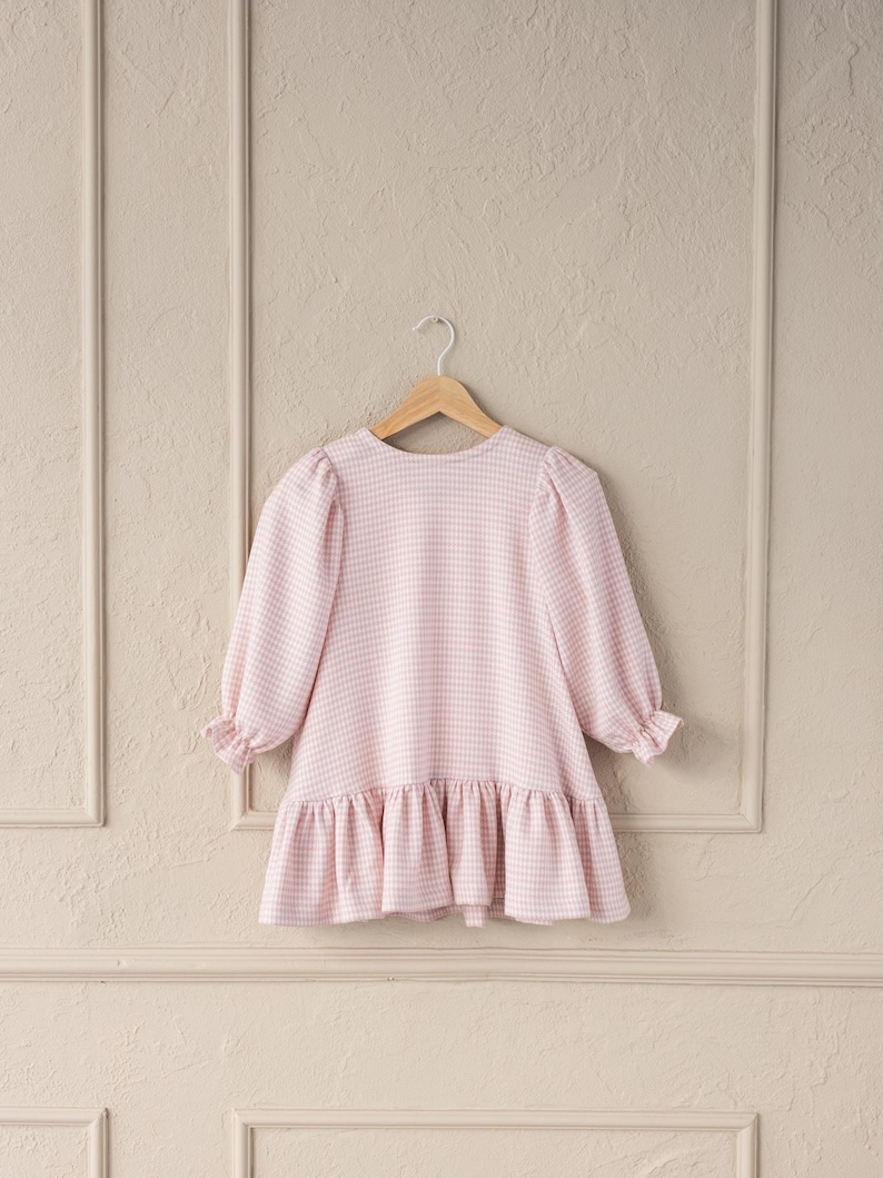 May include: A light pink and white gingham dress with long sleeves and a ruffled hem. The dress is hanging on a wooden hanger against a neutral background. The dress has a round neckline and puffed sleeves.
