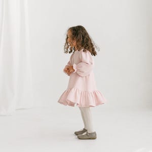 May include: A light pink dress with a textured pattern and a ruffled hem. The dress has long, puffy sleeves and is worn with white tights and gray shoes. The dress is designed for children.