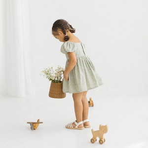 Puede incluir: Una ni&ntilde;a peque&ntilde;a con un vestido verde floral y sandalias blancas sostiene una cesta de mimbre llena de flores blancas. Est&aacute; de pie frente a una pared blanca con una cortina blanca en el fondo. Hay juguetes de madera en el suelo.
