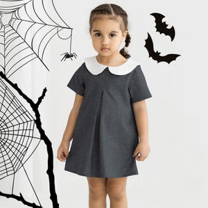 May include: A young girl wearing a dark grey dress with a white Peter Pan collar. She is standing in front of a white background with black spiderwebs and bats.