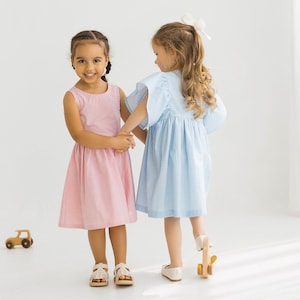 May include: Two young girls wearing matching gingham dresses, one in pink and one in blue, hold hands and smile at the camera. The girl in the pink dress is looking at the camera, while the girl in the blue dress is looking away.