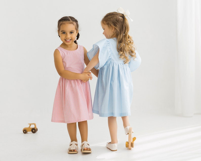 May include: Two young girls wearing matching gingham dresses, one in pink and one in blue, hold hands and smile at the camera. The girl in the pink dress is looking at the camera, while the girl in the blue dress is looking away.