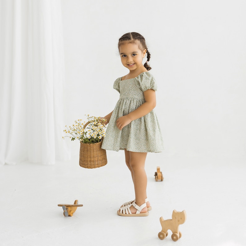 Puede incluir: Una ni&ntilde;a peque&ntilde;a con un vestido verde floral sostiene una cesta de mimbre llena de flores blancas. Lleva sandalias blancas y est&aacute; de pie sobre una superficie blanca. Hay juguetes de madera en el suelo, incluyendo un avi&oacute;n y un gato.