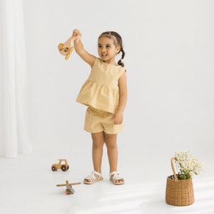 May include: A young girl wearing a yellow and white gingham dress and shorts set. She is holding a wooden toy airplane and smiling at the camera.