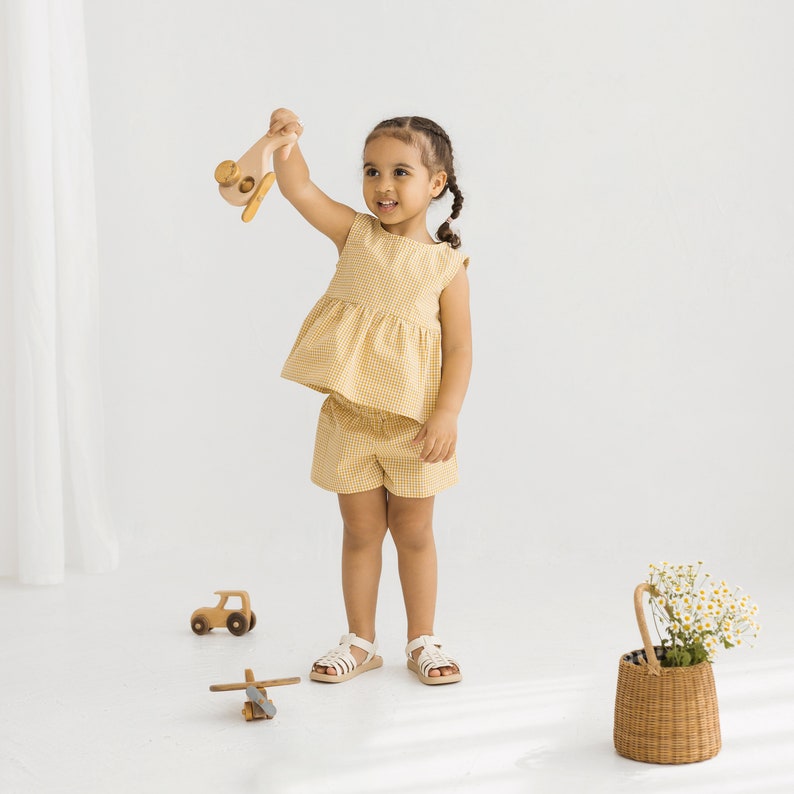 May include: A young girl wearing a yellow and white gingham dress and shorts set. She is holding a wooden toy airplane and smiling at the camera.
