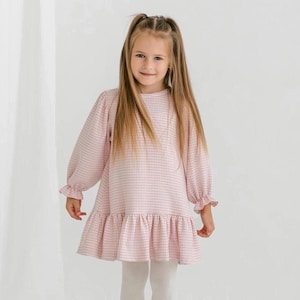 Puede incluir: Un vestido rosa claro y blanco a cuadros con mangas largas y un dobladillo con volantes. El vestido tiene un cuello redondo y se usa con leggings blancos. Las mangas tienen puños con volantes. El vestido lo lleva una niña con pelo largo y rubio.