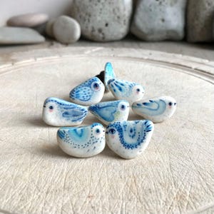 May include: Seven small ceramic bird figurines painted in shades of blue and white. The birds are arranged in a semi-circle on a light wood surface.