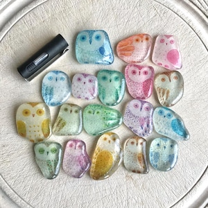 May include: A collection of colorful, glass owl-shaped decorative pieces in various colors, including blue, pink, green, and yellow. The owls have detailed features and are arranged on a white, textured surface. A black lighter is also present.