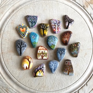 May include: A collection of 15 painted rocks with various designs, including trees, houses, and hearts. The rocks are arranged in a circle on a white surface.