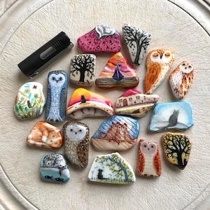 May include: A collection of hand-painted rocks featuring various designs, including owls, trees, landscapes, and cats. The rocks are painted with vibrant colors and intricate details, showcasing a variety of artistic styles and themes.