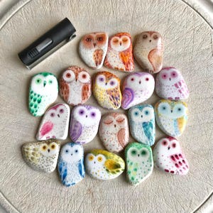 May include: A collection of colorful, hand-painted owl-shaped decorative stones. The owls feature various designs with colors like orange, green, purple, and blue. A black lighter is also present in the image.