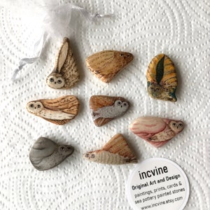 May include: Set of nine hand-painted owl figurines on natural stones. Each owl is painted in a different colour and style, with details like feathers and eyes. The stones are various shapes and sizes. The image includes a business card with the text "incvine Original Art and Design paintings, prints, cards & sea pottery painted stones www.incvine.etsy.com".